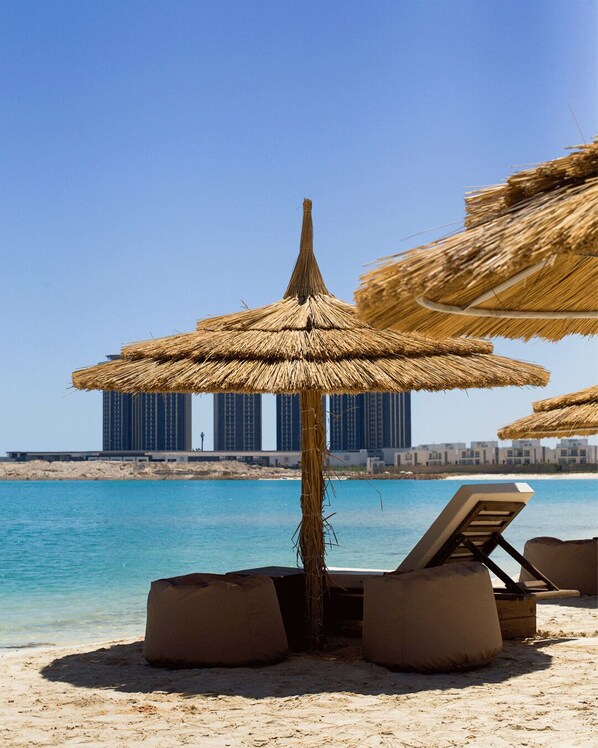 A beach with thatched umbrellas, lounge chairs, and a view of high-rise buildings in the background.