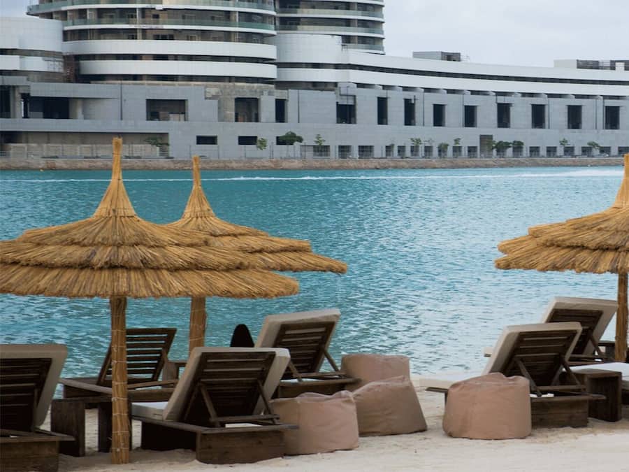 A beach with lounge chairs and thatched umbrellas, a body of water, and a modern building in the background.