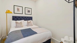 A neatly made bed with a tufted headboard, a bedside lamp, and two framed pictures on the wall. - HS1 - Stylish 3-Bed near Kensington High Street (Greater London)