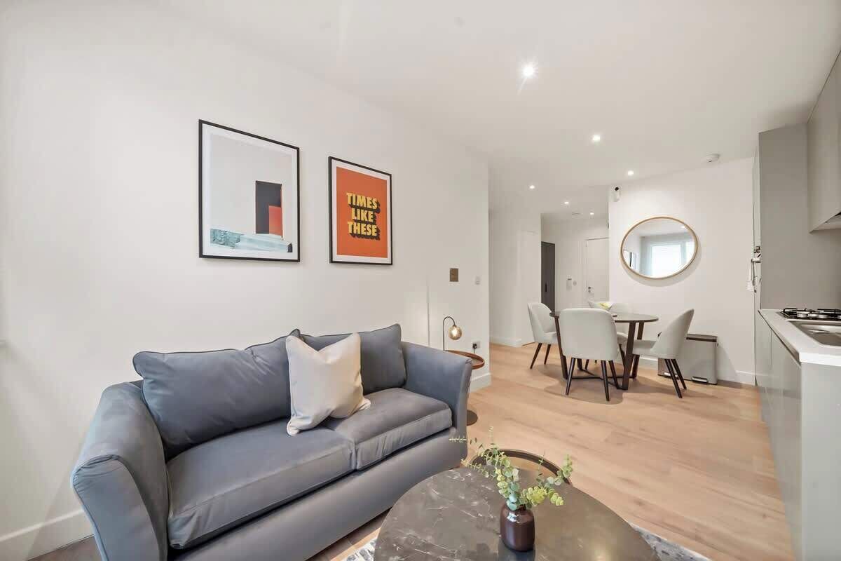 A modern living room with a grey sofa, a coffee table with a vase, a dining area with chairs, and a kitchen with a round mirror.