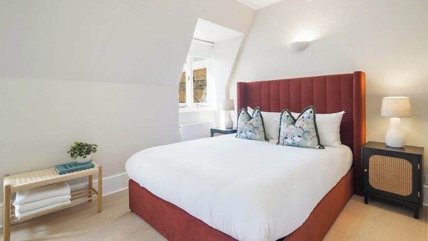 Ein Schlafzimmer mit einem großen Bett, einem Nachttisch mit Lampe, einer kleinen Bank mit Pflanze und einem Fenster mit Blick.