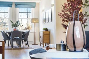 A dining area with a table and chairs, a floor lamp, a rattan cabinet, and a large plant. - HS4 - Beautiful 2-Bed Family Apt in Kensington (Greater London)
