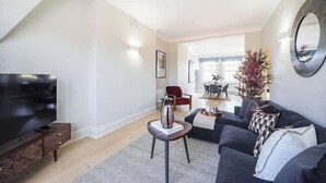 Ein modernes Wohnzimmer mit Flachbildfernseher, dunklem Sofa, Couchtisch und Essbereich im Hintergrund.