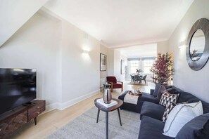 A modern living room with a flat-screen TV, a dark sofa, a coffee table, and a dining area in the background. - HS4 - Beautiful 2-Bed Family Apt in Kensington (Greater London)