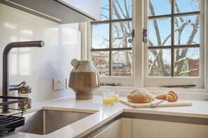 A kitchen with a window, a sink, a mixer, a bottle, and a cutting board with bread and a croissant. - HS4 - Beautiful 2-Bed Family Apt in Kensington (Greater London)