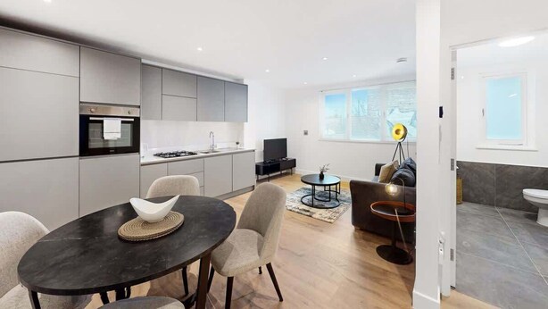 A modern kitchen with grey cabinets, a black countertop, and a dining area with a round table and chairs. - DA1 - Modern & Comfy 1-Bed Boutique Apartment (Greater London)