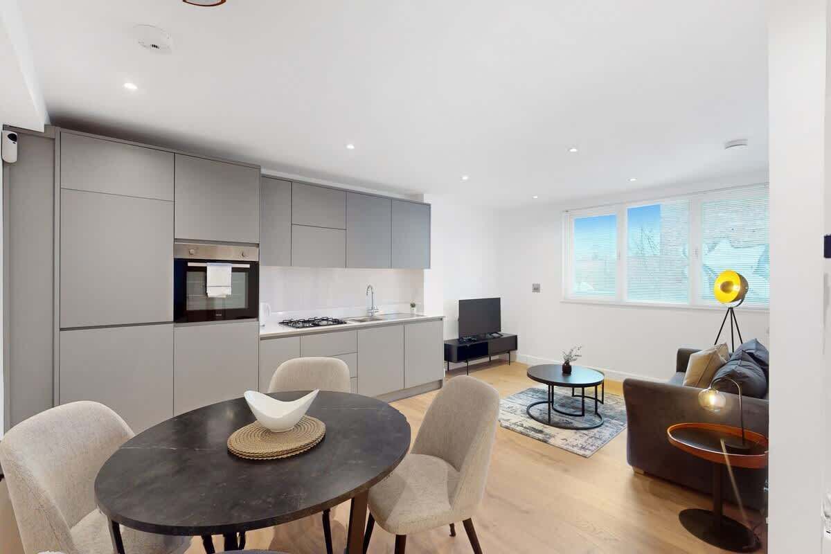 Eine moderne Küche mit grauen Schränken, ein Essbereich mit einem runden Tisch und Stühlen, eine Sitzecke mit Sofa und ein Fernseher in einem Wohnzimmer mit großen Fenstern.