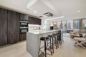 A modern kitchen with a central island, bar stools, and a dining area with a sofa and table. - VW5 - Luxury 3 Bed Townhouse with Parking (Greater London)