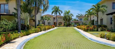 A resort with multiple two-story buildings, a central paved walkway, and well-maintained gardens with palm trees and flowering plants.