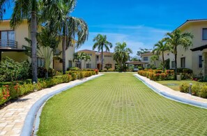 Un resort con varias construcciones de dos pisos, un camino pavimentado central y jardines bien cuidados con palmeras y plantas con flores.