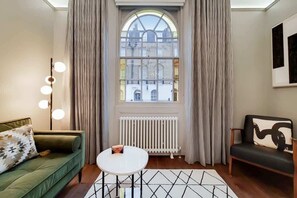 Een kamer met een groene bank, een witte tafel, een zwarte stoel, een radiator en een raam met gordijnen.