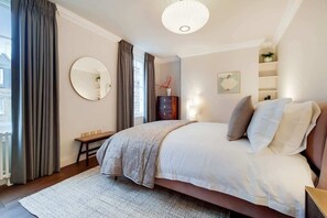 Een slaapkamer met een bed, nachtkastje, spiegel, kaptafel en een raam met gordijnen.