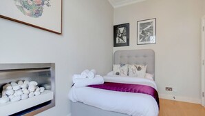 A bedroom with a bed, pillows, a headboard, a wall-mounted shelf with white objects, and two framed pictures. - CG12 - 2 Bedroom Apt, Kensington Garden Square View (London)