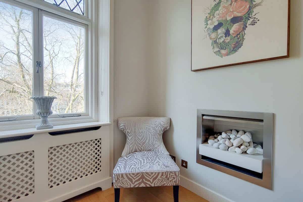 Ein Eckzimmer mit Heizkörper, Stuhl, Kamin und Fenster mit Blick auf Bäume.