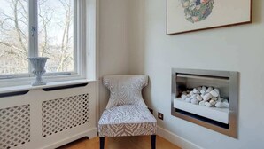A corner room with a radiator, a chair, a fireplace, and a window with a view of trees. - CG12 - 2 Bedroom Apt, Kensington Garden Square View (London)