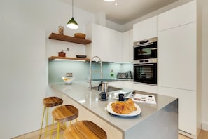 A modern kitchen with white cabinetry, a stainless steel sink, and a built-in oven. There are wooden stools and a breakfast plate with croissants. - BR9 - Elegant 2-Bed near Borough Market (Greater London)