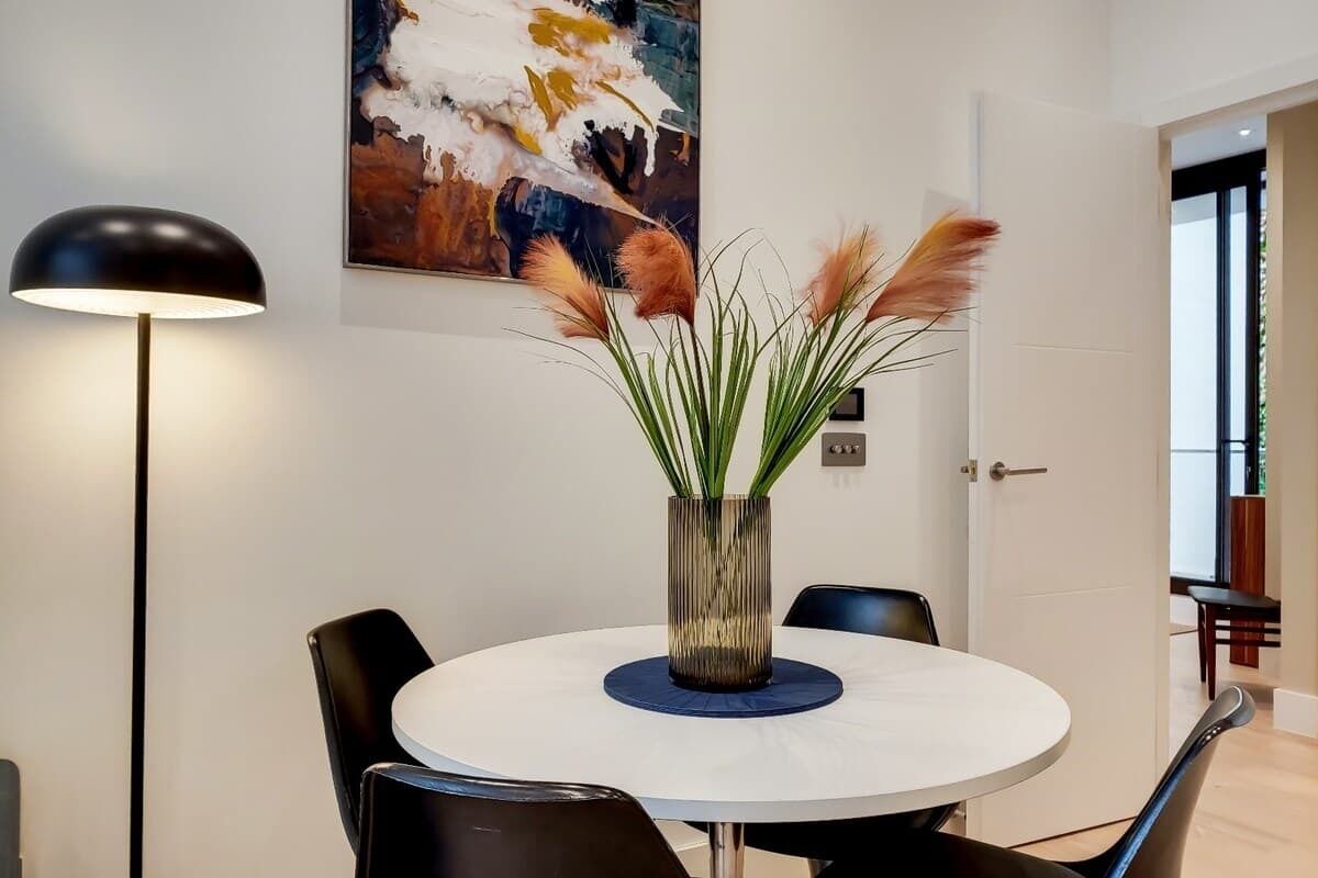 Une table de salle à manger ronde, avec un plateau blanc, est entourée de chaises noires. Un grand vase texturé, contenant des plantes vertes et des plumes d’herbe des pampas, est placé au centre. Une lampe de sol noire se trouve à gauche de la table.
