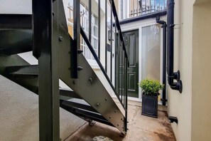 Ein schmaler Außenbereich mit einer Metalltreppe, einem Blumentopf und einer Tür zu einem Gebäude.