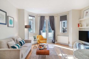 A modern living room with a sofa, coffee table, and a chair. There is a TV mounted on the wall and a window with curtains. - HR2 - Charming 1-Bed near Holland Park (Greater London)