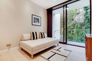 A modern living room with a sofa, a framed picture on the wall, and a view of a garden through large sliding glass doors. - BR11 - Contemporary 2-Bed with City Vibes (Greater London)