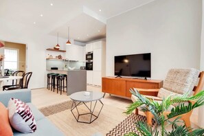 Ein modernes Wohnzimmer mit Sofa, Couchtisch, Fernseher und Essbereich mit Stühlen sowie einer Küche im Hintergrund.