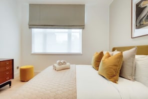 Un dormitorio con cama, almohadas, una mesita de noche, una ventana y un cuadro en la pared.