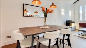 Un comedor moderno con una mesa de madera, sillas blancas, luces colgantes, un jarrón con plantas secas y un espejo grande en la pared.