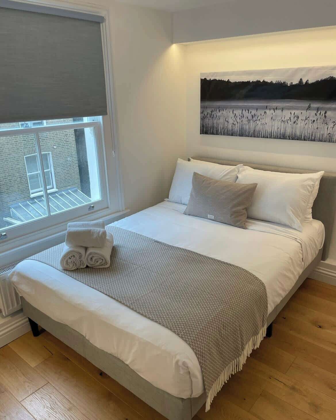 A neatly made bed with white linens and a grey throw, a window with blinds, and a painting of a landscape on the wall.