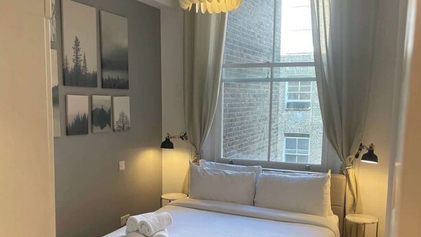 A modern bedroom with a large bed, a hanging light fixture, a window with curtains, and artwork on the wall. - LG4 - Modern & Spacious 2-Bed in Notting Hill (Greater London)