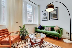 A living room with a green sofa, a brown leather chair, a glass coffee table, and a floor lamp. There are two framed pictures on the wall and a potted plant. - BR8 - Spacious 2-Bed Apartment Near Borough Market (Greater London)