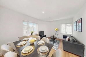 Ein modernes Wohnzimmer mit Esstisch für vier Personen, einer Couch, einer Stehlampe und einem Fernseher.
