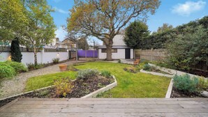 Ein Garten mit einer Holzterrasse, einem großen Baum und einem weißen Gebäude mit einer violetten Wand.
