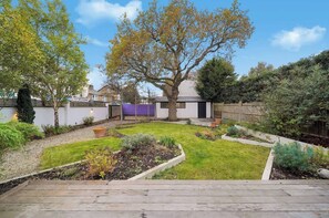 A backyard with a wooden deck, a large tree, and a white building with a purple wall. - MUR1 - Modern and Stylish 6-bed House in Wimbledon (London)