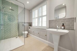 Ein Badezimmer mit Duschkabine aus Glas, Toilette, Waschbecken mit Spiegel und Fenster mit Jalousien.