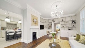 Ein geräumiges Wohnzimmer mit Kamin, einer Chandelierleuchtre und einer modernen Küche mit weißen Schränken.