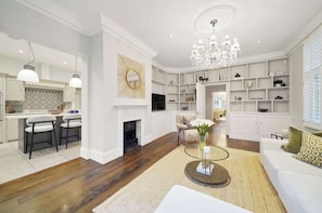 A spacious living room with a fireplace, a chandelier, and a modern kitchen with white cabinetry. - MUR1 - Modern and Stylish 6-bed House in Wimbledon (London)
