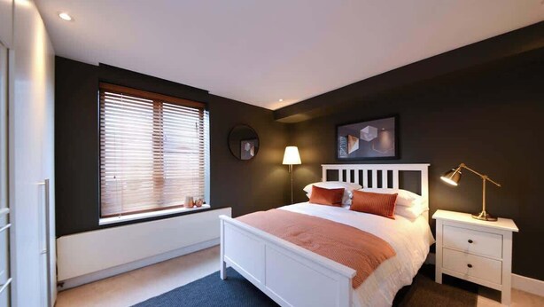 Quarto com cama, mesa de cabeceira, abajur, janela com persianas e relógio de parede.