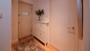 Um corredor com piso de madeira, uma porta branca, um aparador branco com gavetas e uma pequena planta sobre o aparador.