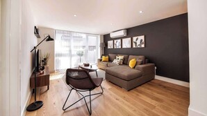 Ein modernes Wohnzimmer mit einer Couch, einem Couchtisch und einem Sessel. Eine Stehlampe, ein Beistelltisch und eine Pflanze sind ebenfalls vorhanden. Der Raum hat Holzböden und ein großes Fenster mit Jalousien.