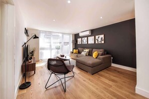 A modern living room with a sofa, a coffee table, and a chair. There is a floor lamp, a side table, and a plant. The room has wooden flooring and a large window with blinds. - BR7 - Modern 2-Bed Apartment with Private Terrace (Greater London)