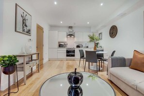 A modern living room with a glass coffee table, a sofa, a dining area with chairs, a kitchen with cabinets and appliances, and a door. - BR2 - Stunning 1 Bed Apartment Heart of Borough (Greater London)