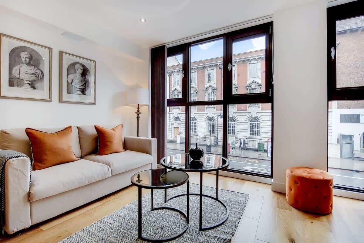 A modern living room with a sofa, two side tables, a red ottoman, and large windows with a city view.