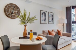 Ein modernes Wohnzimmer mit einem runden Esstisch, einer Couch mit Kissen, einer großen Wanduhr, gerahmten Kunstwerken und einem Topf mit Pflanze.