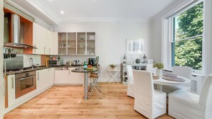 Une cuisine moderne avec des meubles blancs, un coin repas avec des chaises blanches et une grande fenêtre donnant sur les arbres.