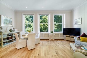 A living room with a large TV, white furniture, a bookshelf, and large windows with a view of trees. - CG9 - 1 Bedroom Apt, Kensington Garden Square View (Greater London)