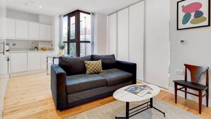 Una sala de estar moderna con un sofá, una mesa de centro y un sillón. Hay una zona de cocina con armarios blancos y una ventana.
