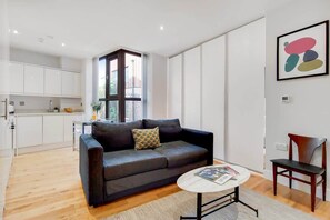 Ein modernes Wohnzimmer mit einer Couch, einem Couchtisch und einem Sessel. Es gibt eine Küchenzeile mit weißen Schränken und einem Fenster.