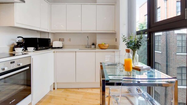 Una cocina moderna con armarios blancos, un horno Bosch, un microondas negro y una mesa de vidrio con una planta y un vaso de jugo de naranja.
