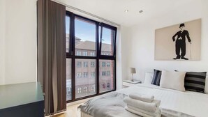 Habitación de hotel con un ventanal grande, una cama con sábanas blancas, una mesita de noche y un cuadro en la pared.