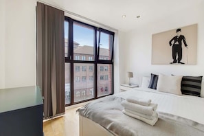 A hotel room with a large window, a bed with white linens, a nightstand, and a painting on the wall. - BR6 - Beautiful 1 Bed Flat in Heart of Borough (Greater London)